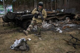 Украинский военный пинает сгоревший спальный мешок. На фоне — сгоревший российский танк. Стоянка, Украина