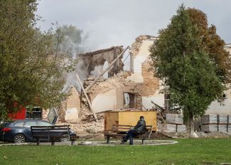 Мужчина сидит на скамейке у поврежденного здания 