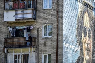Мужчина сидит на балконе дома, на стене которого нарисовано граффити с музыкантом Джоном Леноном