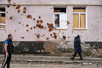 60-летний Андрей Плешан (слева) стоит у изрешеченной стены дома в Изюме. Во время российской оккупации города — с апреля по сентябрь 2022 года — Плешан укрыл от обстрелов до 60 человек в своем подвале