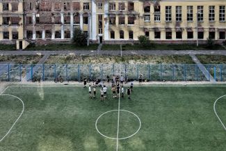 Подростки играют в футбол перед зданием школы, поврежденным в результате военных действий