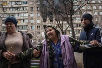Люди помогают пожилой женщине пройти по улице мимо обстрелянного дома. 7 марта 2022 года