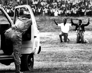 Юноши из Соуэто стоят на коленях с поднятыми руками перед целящимся в них полицейским. В июне 1976 года молодежь Соуэто, гетто для чернокожих, взбунтовалась против школьной реформы, которая предписывала перевести обучение на африкаанс вместо английского и языков коренных народов Африки. Полиция жестоко подавила эти выступления, открыв огонь по безоружным демонстрантам. Многие сотни из них погибли. Соуэто, ЮАР, июнь 1976 года