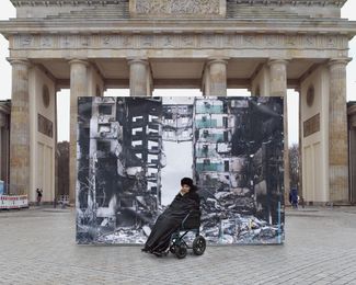 Olena, a 92-year-old former teacher, against the backdrop of a photograph showing a destroyed apartment building in Borodyanka, Kyiv region. The photograph, taken by Maxim Dondyuk, was placed in front of the Brandenburg Gate in Berlin, Germany.