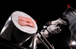 Симон Делестр из сборной Франции по верховой езде на фоне барьера в виде гигантского традиционного японского ролла с лососем. Снято во время квалификационного заезда на 14-й день Олимпиады.