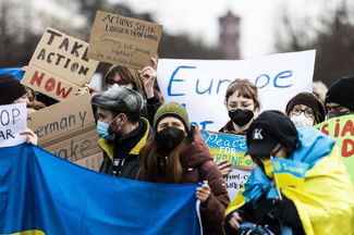 Люди с плакатами и флагами Украины собрались перед Бранденбургскими воротами. Меньше суток назад одна из главных достопримечательностей Берлина была подсвечена голубым и желтым