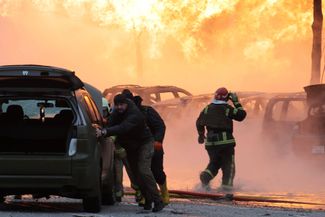 Мужчины пытаются отогнать автомобили от места возгорания газопровода, получившего повреждения в результате падения ракетных обломков в Подольком районе Киева.