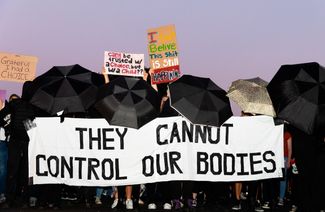 Демонстранты в черной одежде с черными зонтиками в руках проходят мимо Капитолия США. Активисты несут баннер со словами «Они не могут контролировать наши тела»