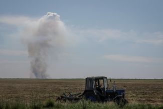 Фермер в Херсонской области управляет трактором. На горизонте виден дым от очередного ракетного удара