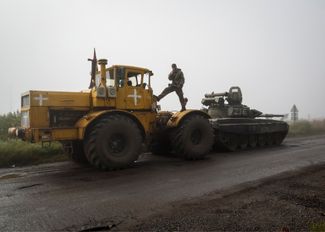Украинский военный стоит на тракторе, который тащит российский танк