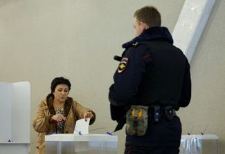 Жительница Москвы голосует на избирательном участке, пока рядом с урной стоит полицейский. 17 марта 2024 года