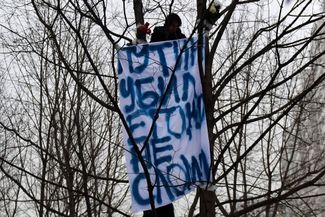 A mourner in a tree holds a sign outside the cemetery that says “Putin killed him but didn’t break him.”